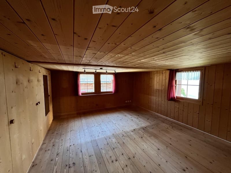 Empty room with wooden walls, wooden floor, and wooden ceiling. Two windows with red curtains, track lighting, and a small mirror on the left wall.