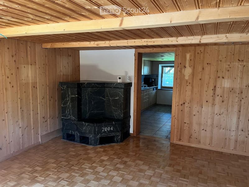 A room with wooden walls and ceiling, featuring a black marble fireplace with the year 2004 inscribed on it. The room has a wooden floor and an open doorway leading to a kitchen area with cabinets and appliances.