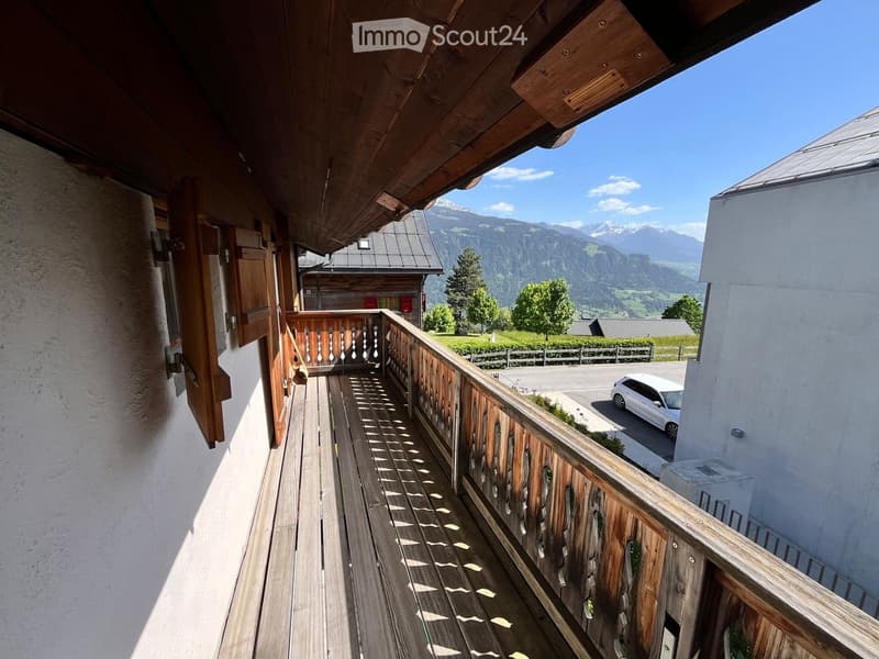 Wooden balcony with railing, overlooking a scenic mountain view and a parking area with a white car.