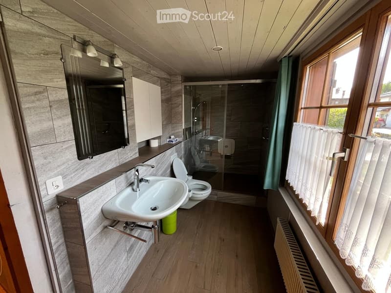 Modern bathroom with a sink, toilet, mirror, and glass-enclosed shower area. Features wooden flooring, grey tiled walls, and a window with white curtains.