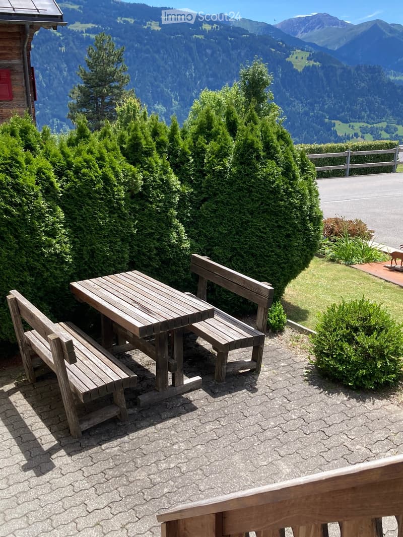 Outdoor seating area with a wooden table and benches, surrounded by neatly trimmed bushes and trees, overlooking a scenic mountain landscape.