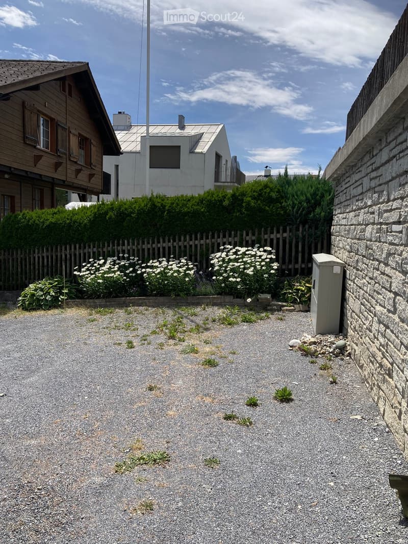Gravel driveway bordered by a wooden fence, with white flowers and shrubs, and a stone wall on one side.