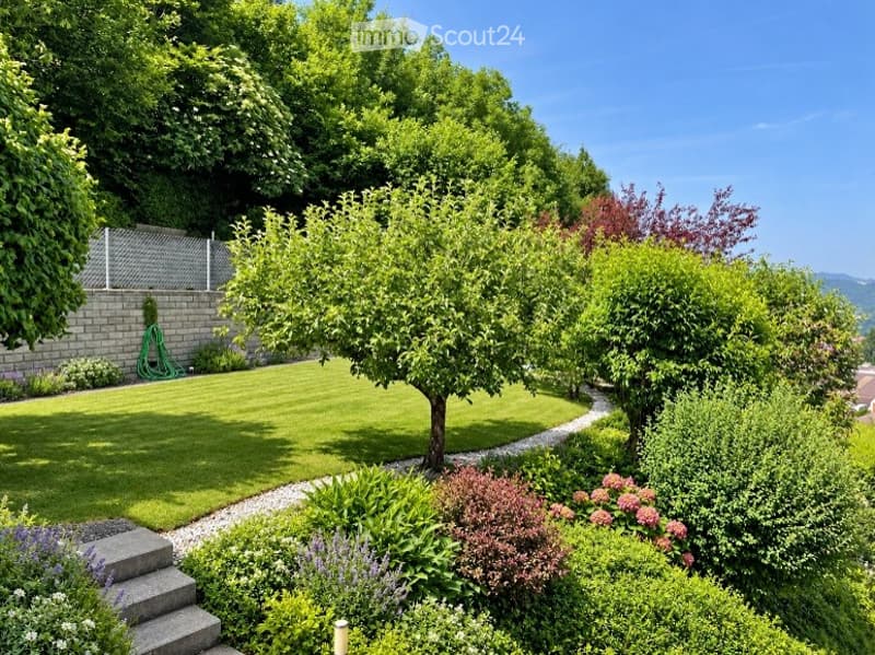 Well-maintained garden with a neatly trimmed lawn, various trees, shrubs, and flowering plants. A gravel pathway winds through the garden, and a small staircase leads up to a higher level.