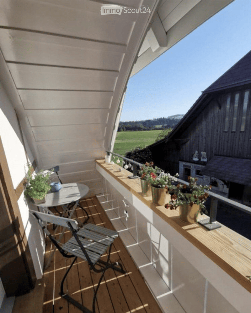 Balcony with wooden decking, small table, chairs, and potted plants, overlooking a green field and mountains.