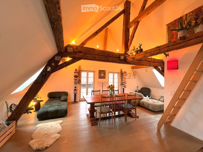 Cozy attic living space with exposed wooden beams, a green sofa, a dining table with chairs, an ottoman, and a bookshelf. The room features warm lighting, a white fur rug, and large windows letting in natural light.
