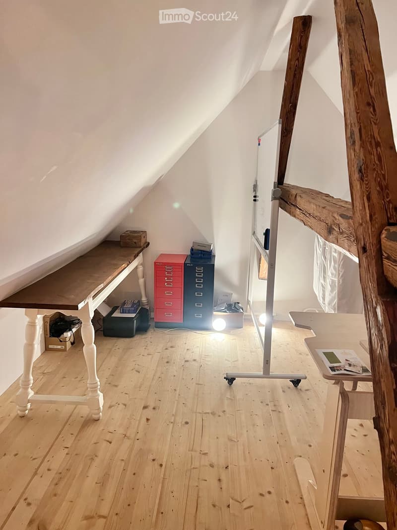 A small attic room with wooden flooring, slanted ceilings, and a large wooden table with a white chair. There is a red and blue filing cabinet, a small stool, and a lamp on the floor.