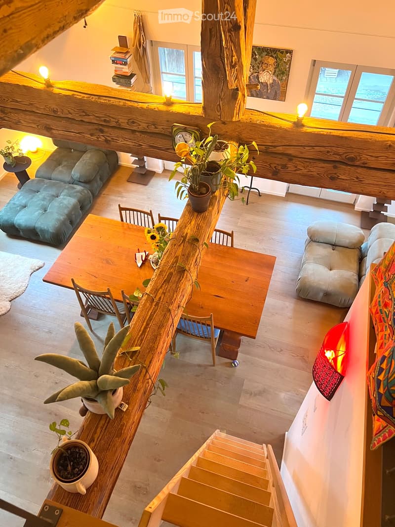 Open-concept living and dining area with wooden floors, large windows, and a staircase leading to an upper level. Features include a sofa, dining table with chairs, potted plants, and decorative lamps.