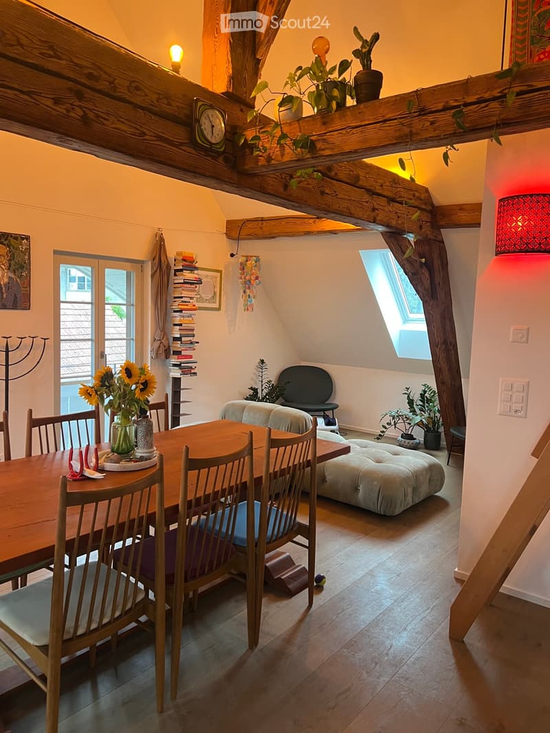 Cozy attic room with wooden beams, skylight, dining table with chairs, sofa, ottoman, potted plants, and bookshelves.