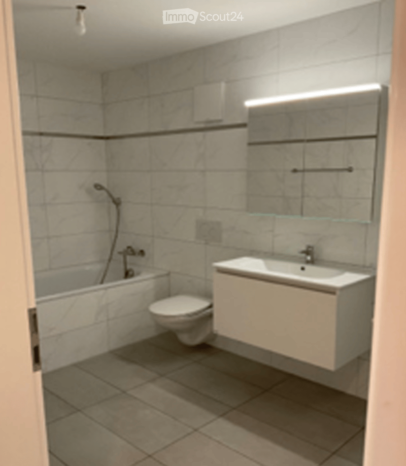 Modern bathroom with a bathtub, toilet, sink, and a large mirror with built-in lighting. The walls are tiled with a mix of light and dark tiles, and the floor is covered with beige tiles.