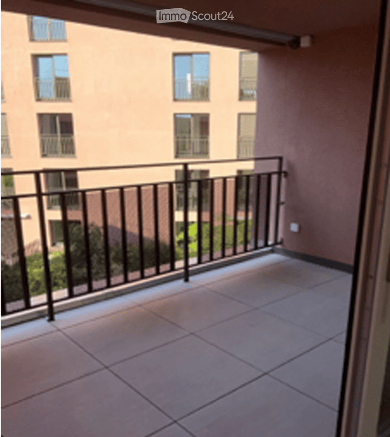 Balcony with tiled floor and black metal railing, overlooking a multi-story building with balconies and windows.