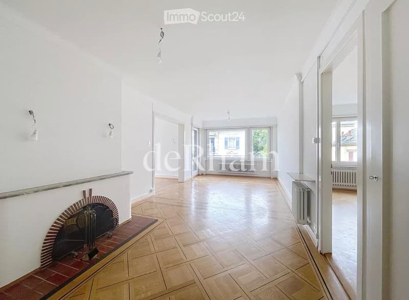 Bright hallway with wooden flooring, white walls, and a fireplace on the left side. The hallway leads to an open space with large windows and radiators visible in the background.