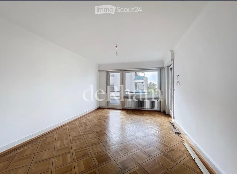 Empty room with wooden parquet flooring, white walls, large window with balcony access, radiator, and a light fixture on the ceiling.