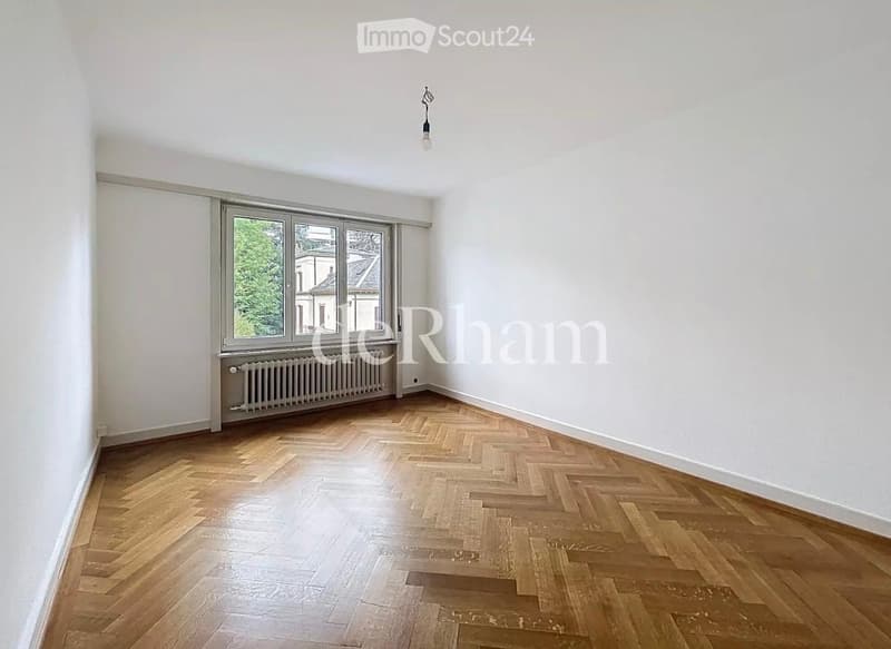 Empty room with wooden herringbone flooring, white walls, large window with radiator below, and a single ceiling light fixture.