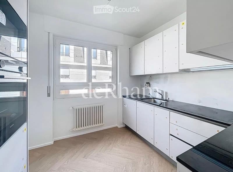 Modern kitchen with white cabinets, a sink, a faucet, and a radiator. The space features wooden flooring and large windows offering a view of the street.