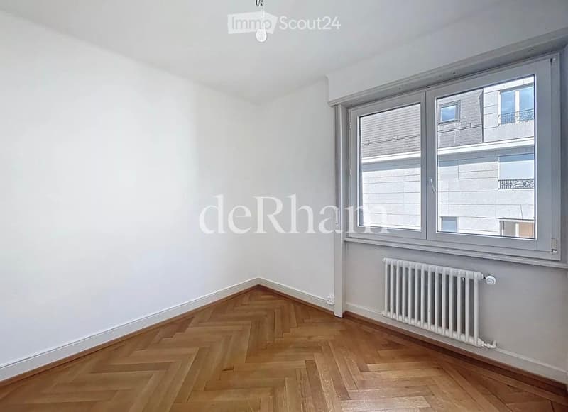 Empty room with wooden herringbone flooring, white walls, large window with blinds, radiator, and a single ceiling light fixture.