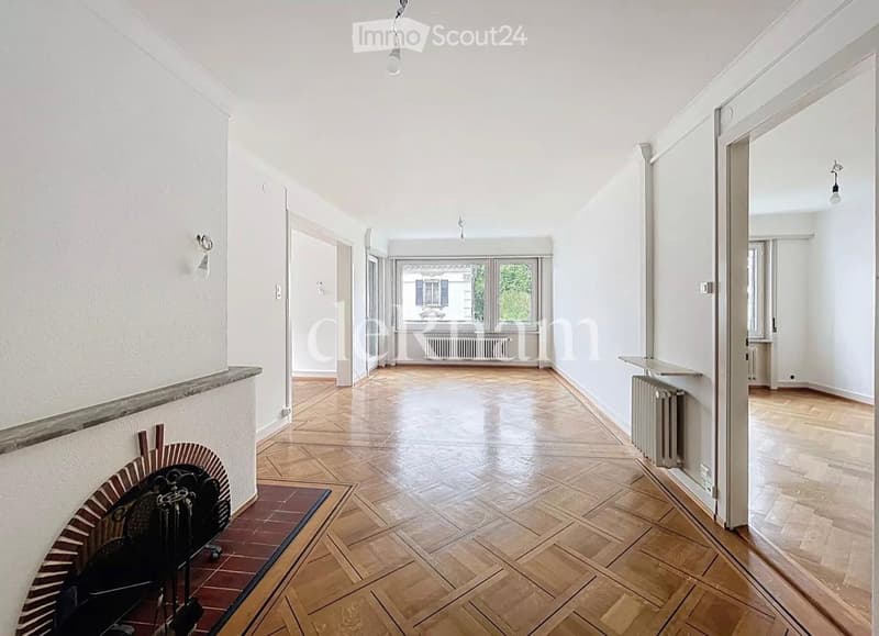 Empty hallway with wooden flooring, white walls, and several doors leading to other rooms. Natural light streams in from a large window at the end of the hallway.