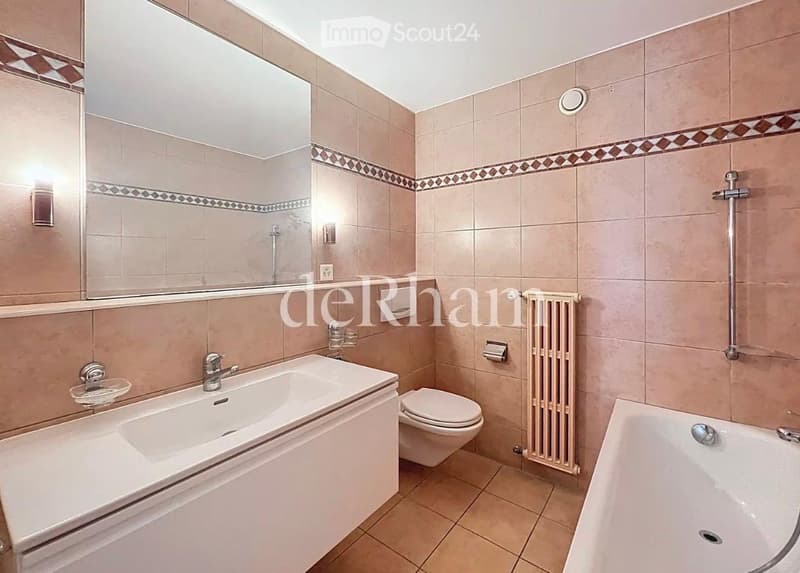 Modern bathroom with beige tiles, a large mirror, a double sink, a toilet, a bathtub, and a radiator.