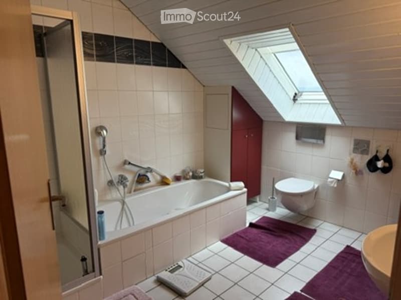 Bathroom with a bathtub, toilet, shower, and a scale on the floor. The room features white tiled walls and floor, with purple towels placed on the floor.