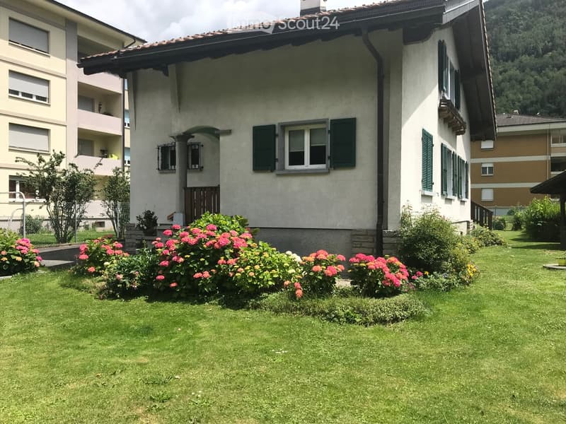 Lush garden with vibrant pink and orange flowers, green grass, and a well-maintained lawn in front of a two-story residential building with white walls, green shutters, and a tiled roof.