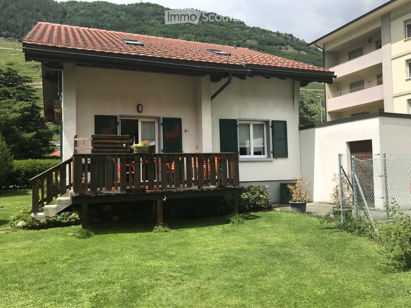 Two-story house with a red-tiled roof, white walls, green shutters, and a wooden balcony with outdoor furniture.