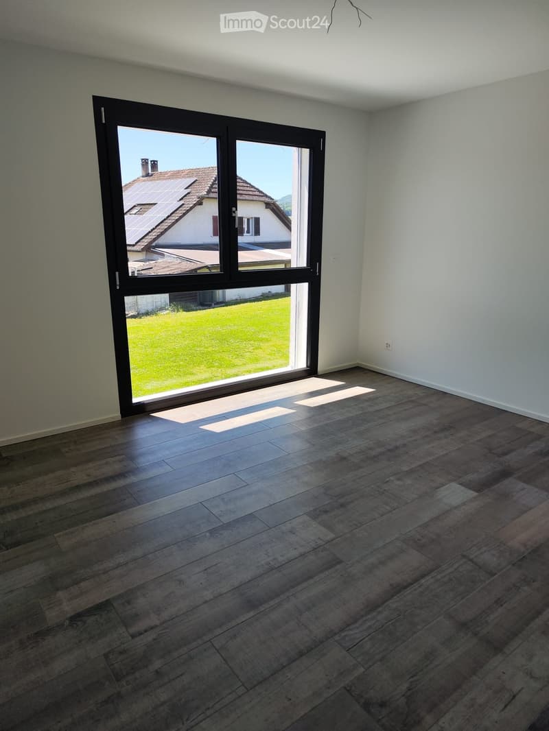 Empty room with wooden flooring and large sliding glass doors leading to a grassy outdoor area and neighboring house with solar panels on the roof.