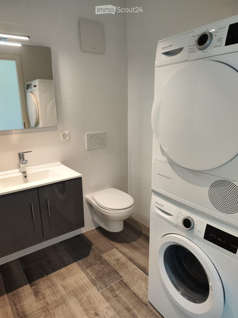 Modern bathroom with a sink, toilet, washing machine, and condenser, featuring dark wooden flooring and white tiled walls.