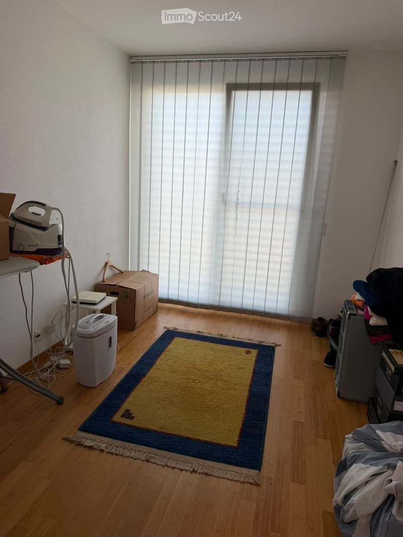 A furnished room with wooden flooring, a large window with vertical blinds, a rug, an ironing board, a printer, a small table, and some boxes.