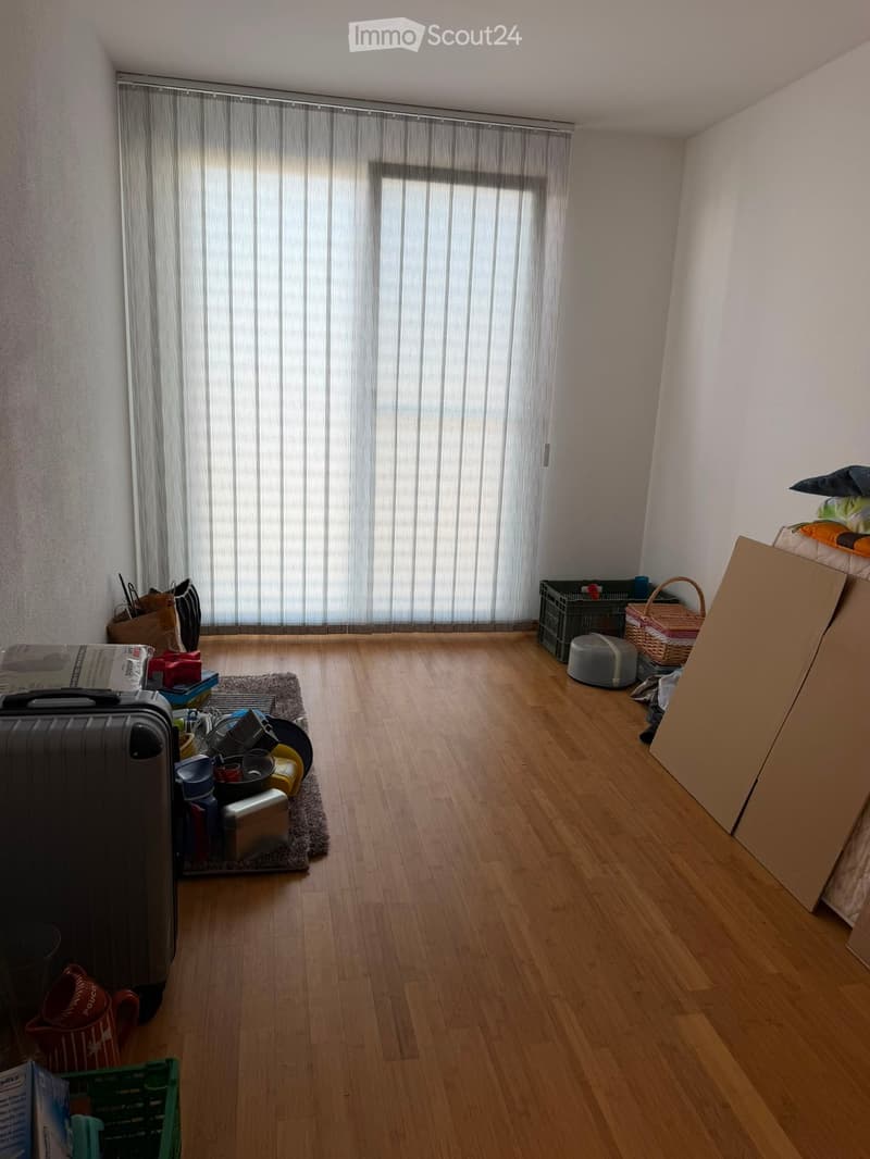 The room has wooden flooring, white walls, and large windows with vertical blinds. Various items are scattered around, including a suitcase, cups, a basket, and some cushions.