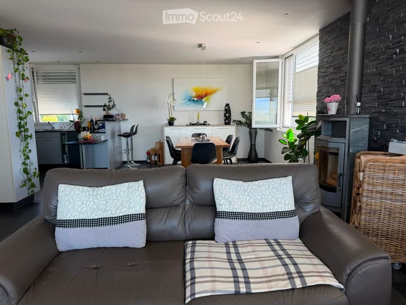 Modern living room with a dark leather sofa, coffee table, dining area, kitchen, and fireplace. Features large windows, plants, and artwork.