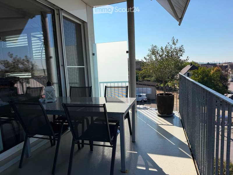Balcony with a glass table, four chairs, a lantern, and a potted plant, overlooking a residential area with trees and a clear blue sky.