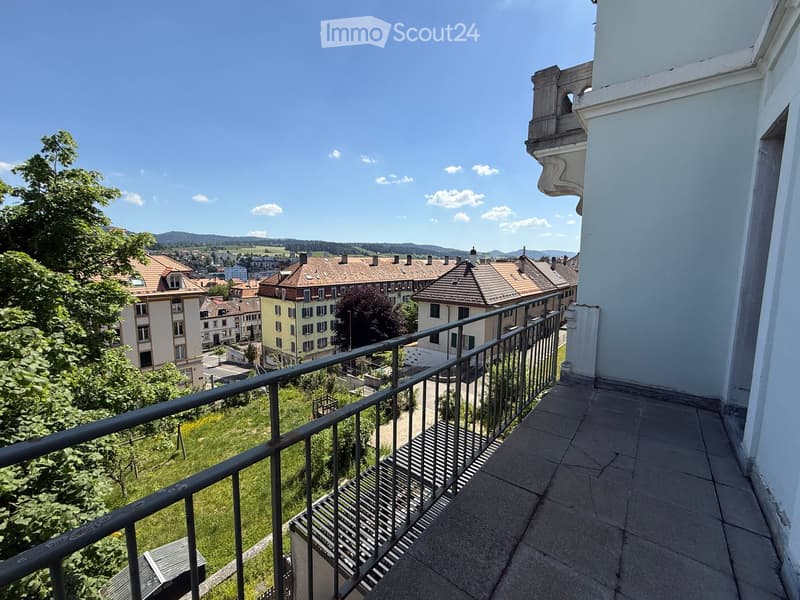 Balcony with metal railing, overlooking a town with houses, trees, and distant hills under a clear blue sky.