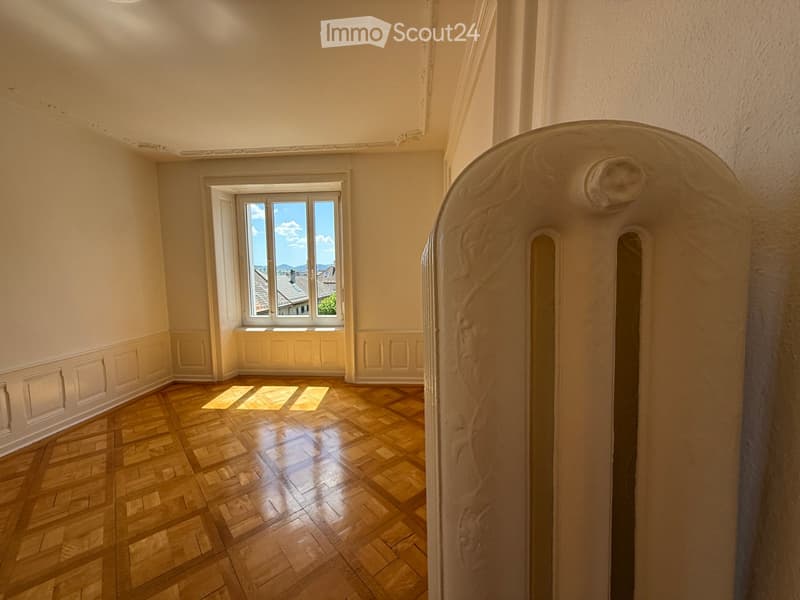 Empty room with wooden flooring, white walls, and a large window offering a view of rooftops and mountains.