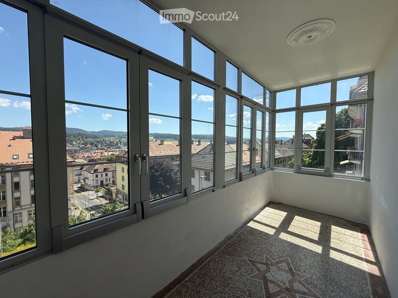 Empty room with large windows offering a view of the cityscape and surrounding landscape. The floor features a patterned tile design, and the walls are painted a light color.