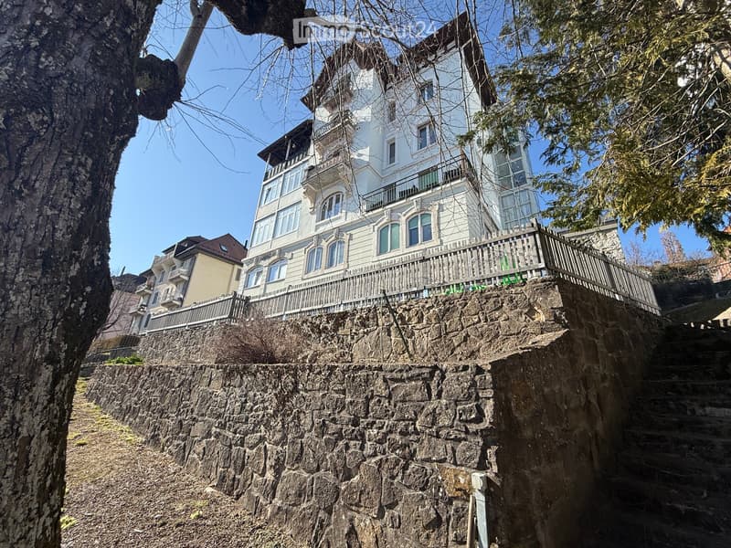 A multi-story residential building with white facade, multiple windows, and balconies, situated on a raised ground floor with stone steps leading up to it.