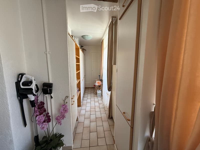 A narrow hallway with tiled flooring, white walls, and a light fixture on the ceiling. There are shelves on the left side and a door at the end of the hallway.