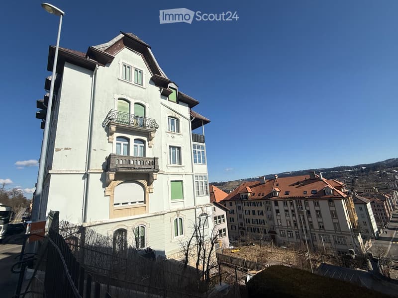 Multi-story residential building with white facade, green shutters, and ornate balconies. The building features a classic architectural style with decorative stonework and multiple windows.