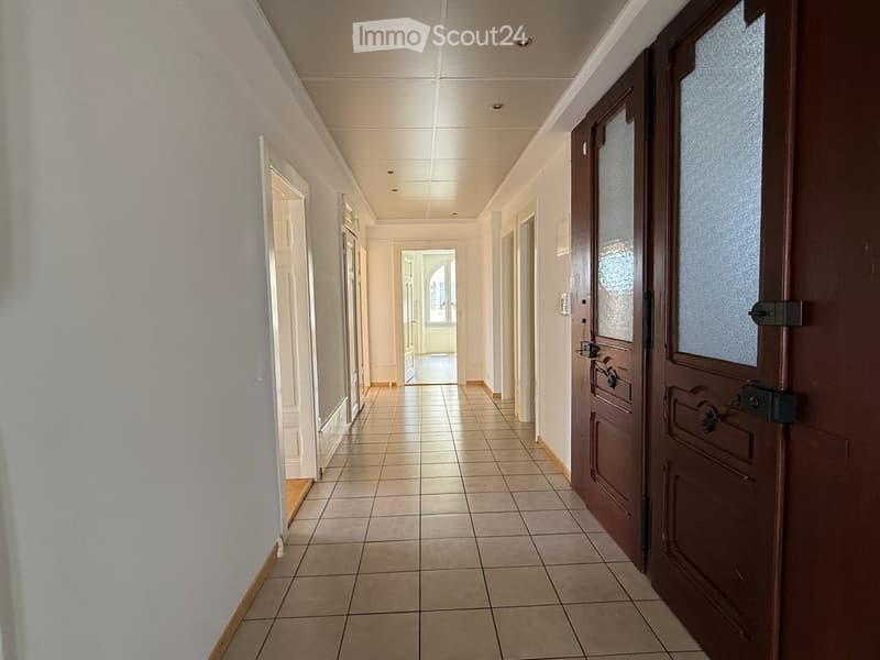 A long, tiled hallway with white walls, multiple doors, and recessed ceiling lighting. The hallway features a polished tile floor and a warm, neutral color palette.