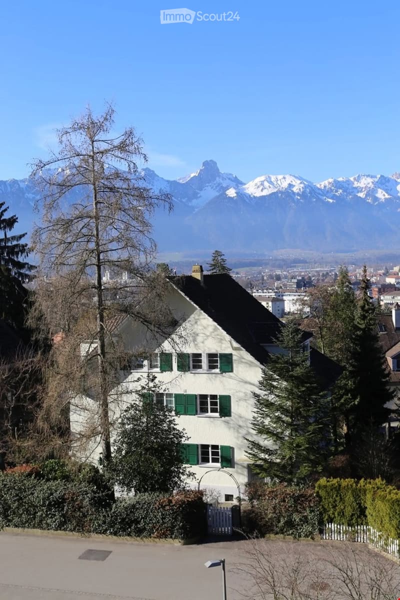 A two-story white house with green shutters, surrounded by trees and bushes, with a scenic mountain view in the background.
