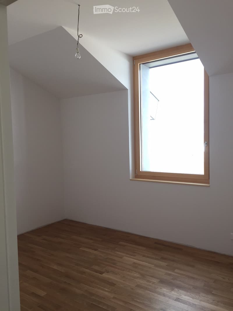 Empty attic room with wooden flooring, white walls, and a skylight window with wooden frame.