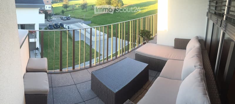 Balcony with a view of a green landscape, featuring a wicker sofa, a coffee table, and two stools.