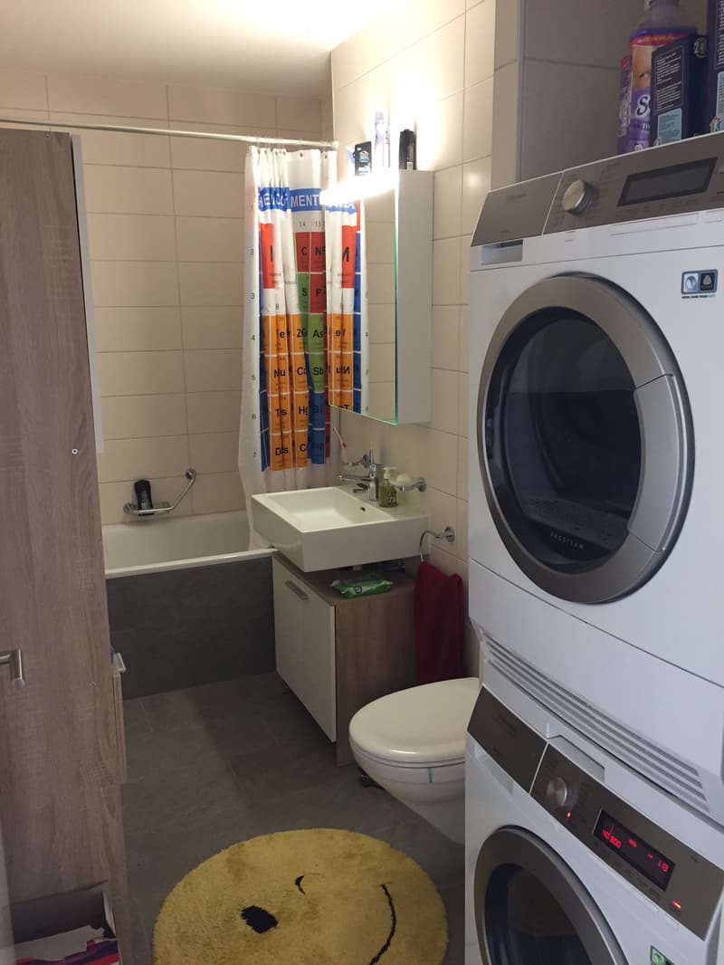 Modern bathroom with a sink, toilet, bathtub, shower curtain, washing machine, and a yellow smiley face rug.
