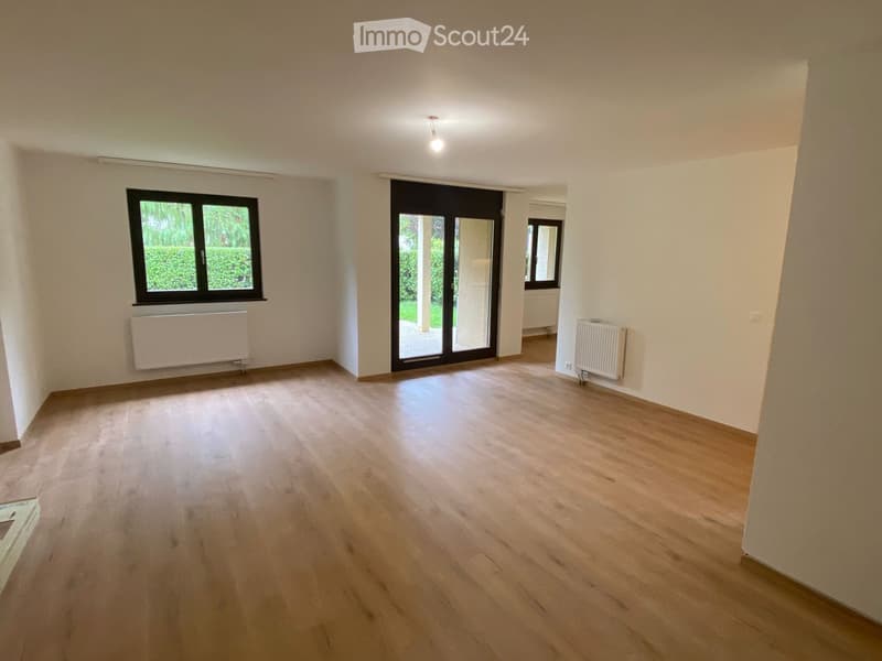 Empty room with light wooden flooring, white walls, and large windows. Features a sliding glass door leading outside and radiators on the walls.