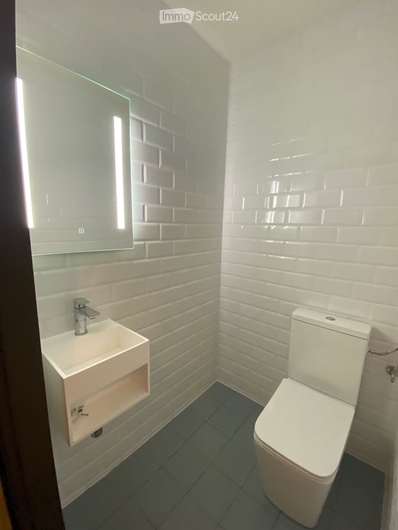Modern bathroom with white brick walls, a white sink with a chrome faucet, a toilet, and a large mirror with built-in lighting.