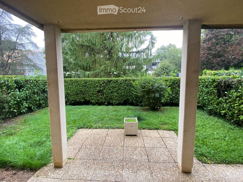 A covered outdoor area with tiled flooring, surrounded by neatly trimmed hedges and a small potted plant in the center.