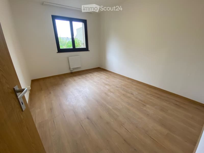 Empty room with wooden flooring, white walls, a window with black frame, and a radiator below the window.