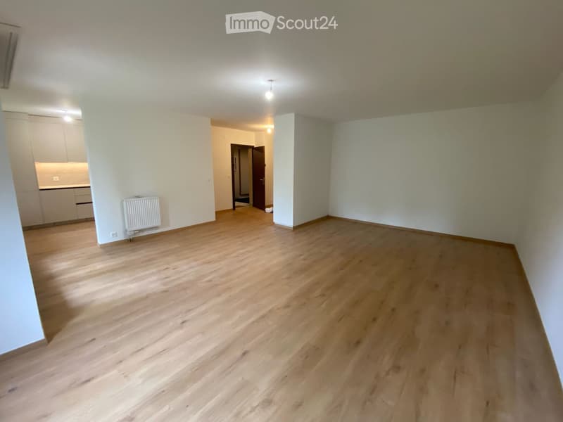 Empty room with wooden flooring, white walls, and a radiator on one wall. The room is connected to a kitchen area with cabinets and a countertop.