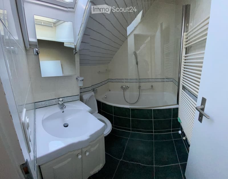 Modern bathroom with a white sink, toilet, bathtub, and mirror. Features dark green tiles and a radiator on the right side.