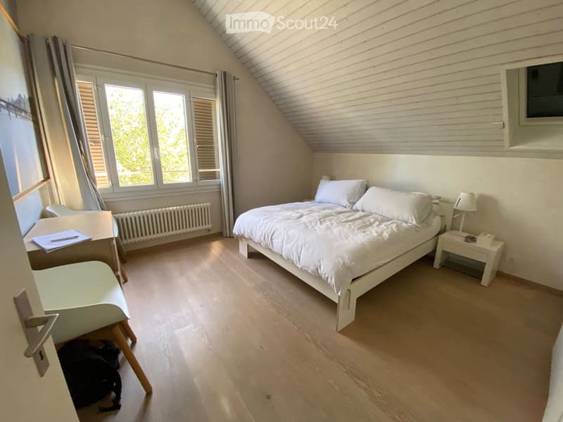 Bright bedroom with wooden flooring, large windows, a double bed with white bedding, a nightstand with a lamp, a desk with a chair, and a radiator under the windows.