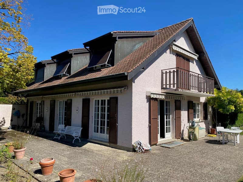 Two-story house with a tiled roof, white exterior, and multiple windows with shutters. Features a balcony on the upper level and a patio area with outdoor furniture and potted plants.