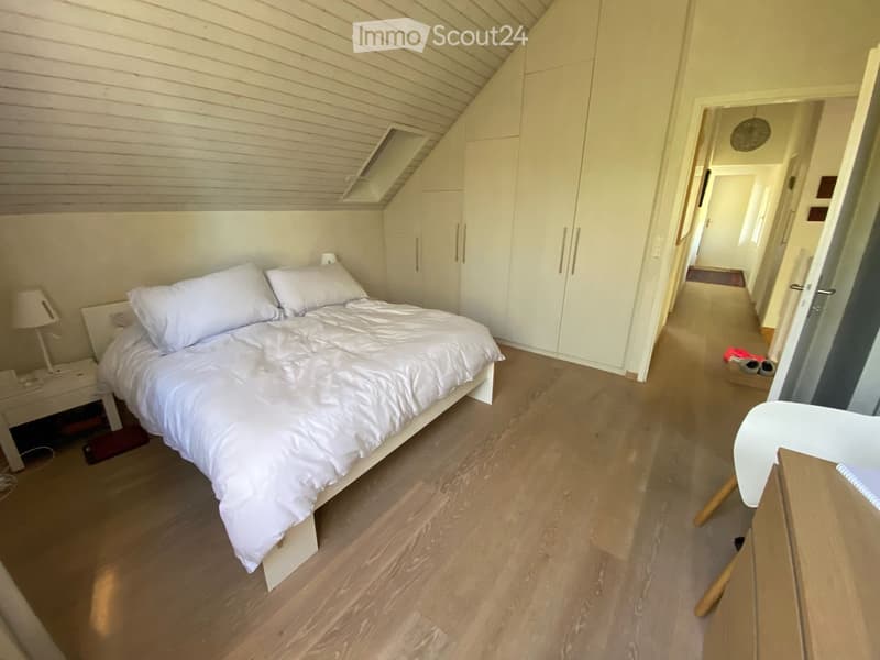 Cozy bedroom with a white bed, nightstand, lamp, and built-in wardrobes. The room features wooden flooring and a slanted ceiling with a skylight.